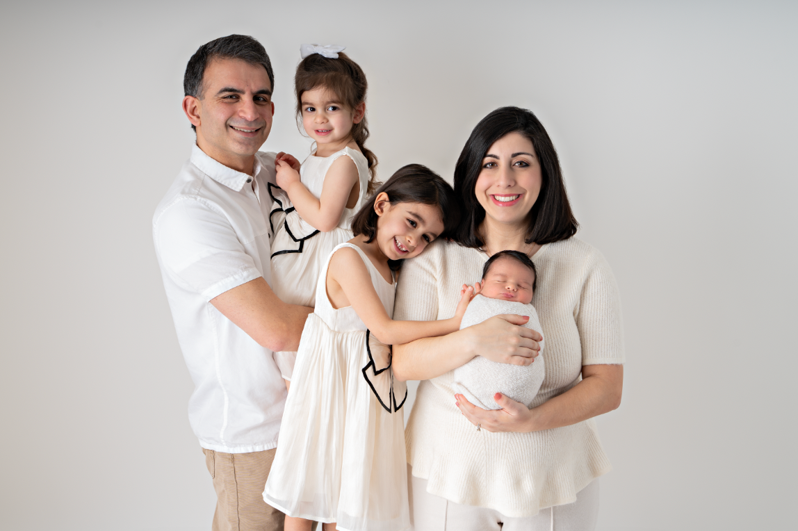 Family of five posing in white outfits, parents holding three young children including a newborn baby, studio portrait.