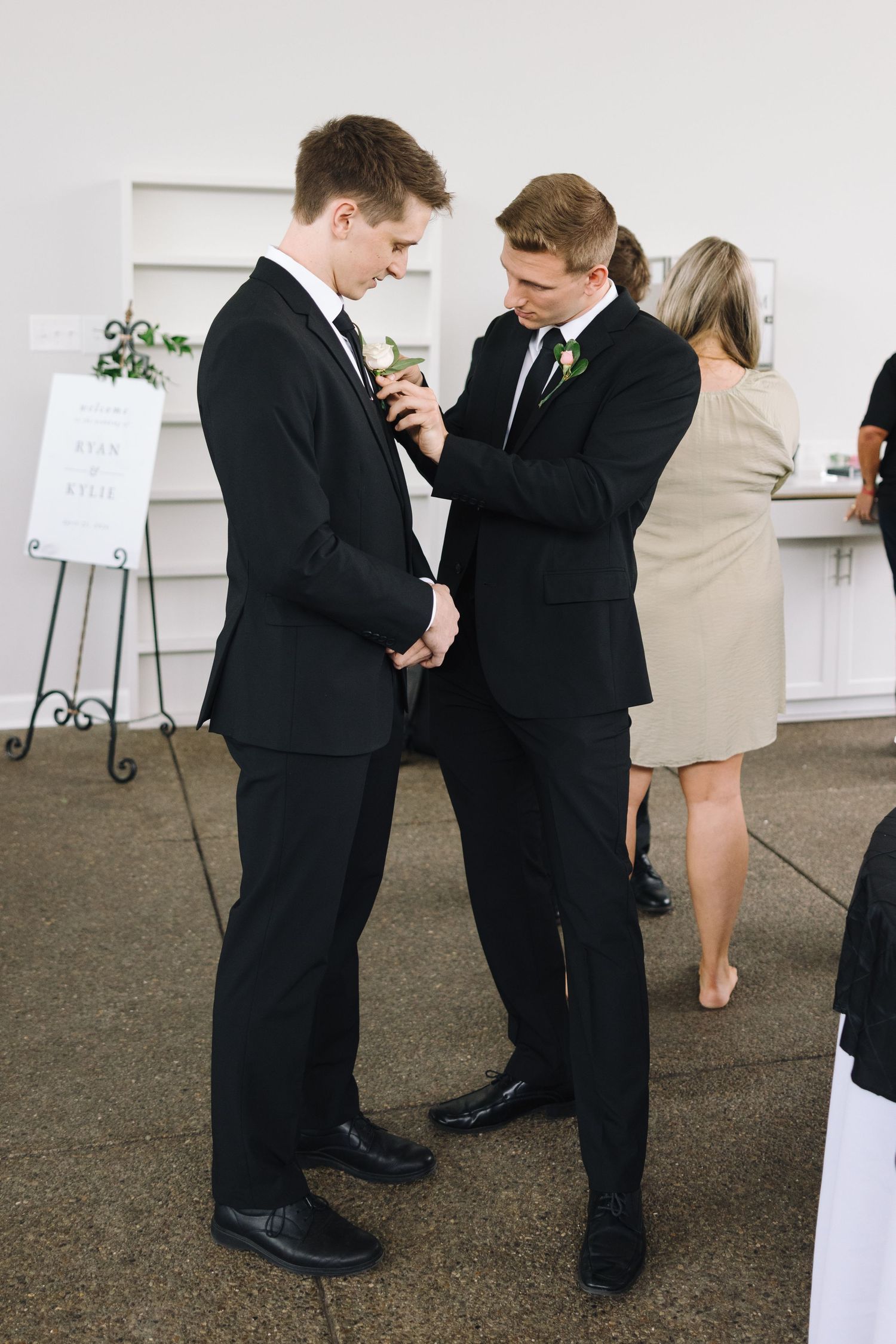 Two people in black suits helping each other with boutonniere pins at wedding.