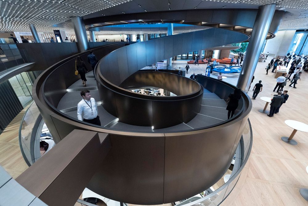 Modern office atrium featuring a spiral staircase with dark metal railings and open-plan workspace with people walking below.