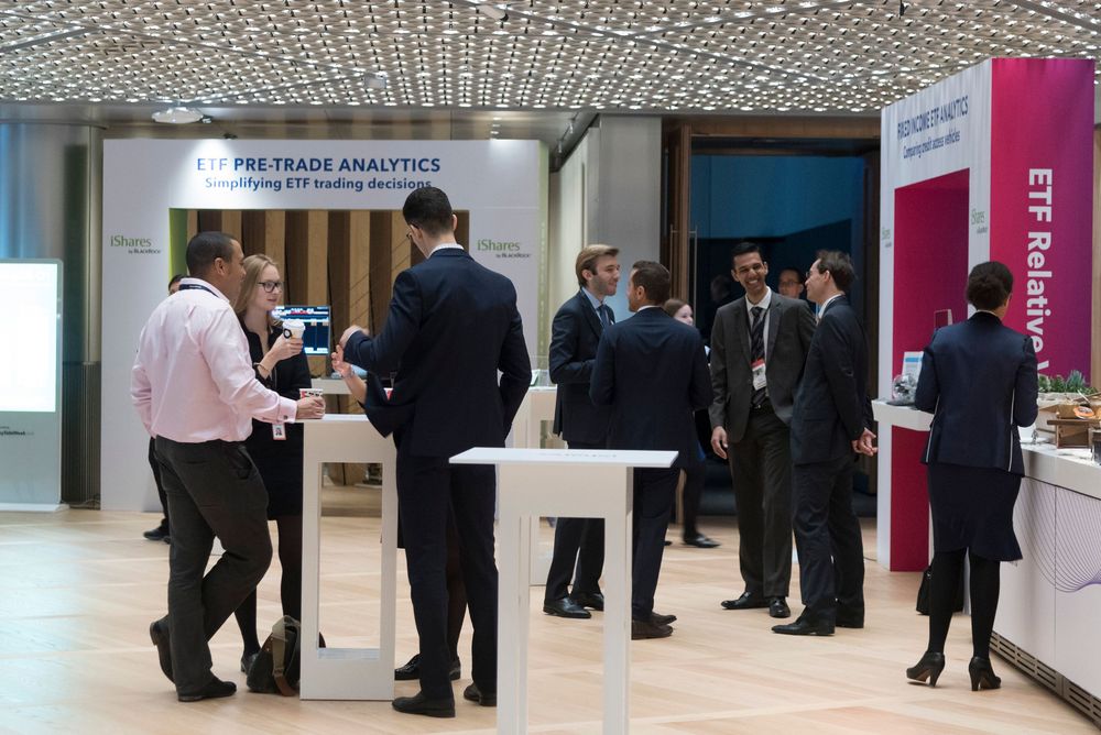 Business professionals networking at an ETF pre-trade analytics conference exhibition booth in a modern venue.