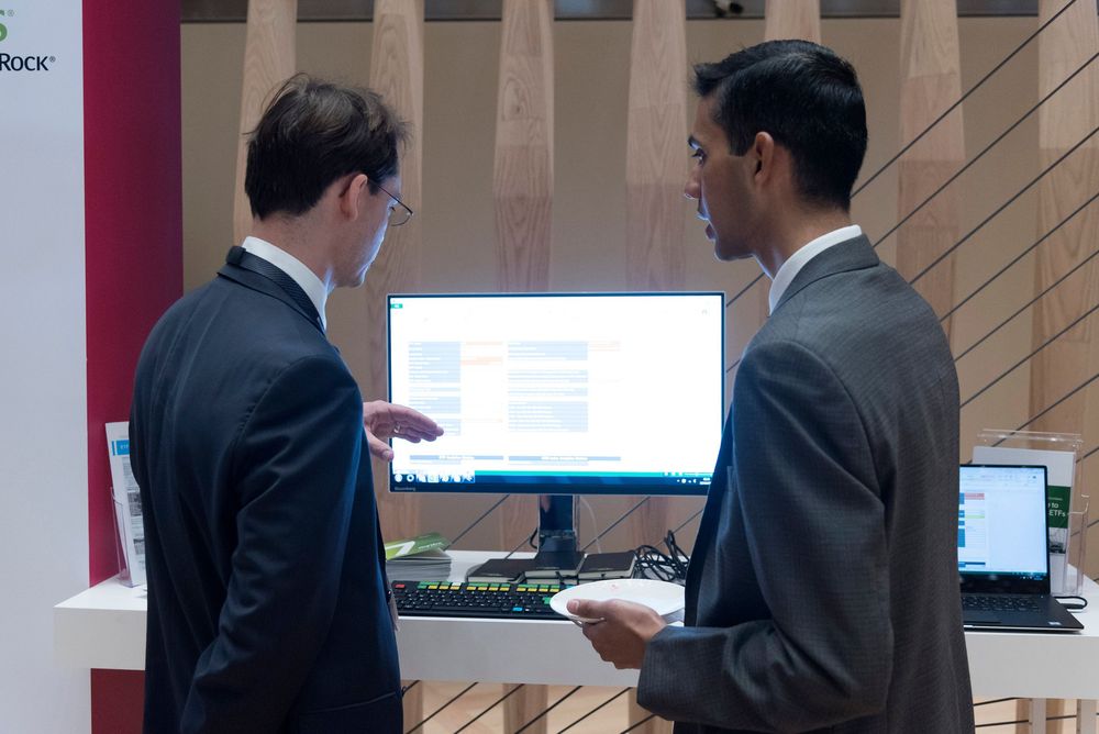 Two businessmen in suits discussing data on a computer monitor at a corporate trade show booth.