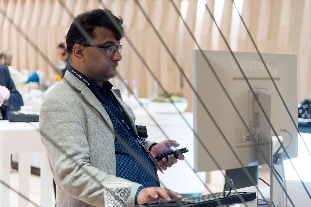 A man in a grey blazer uses a smartphone at a conference podium with technology displays in the background.