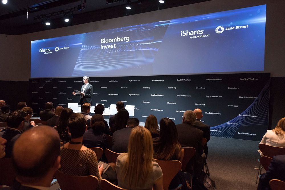 Speaker presents at Bloomberg Invest conference with iShares by BlackRock branding on large screen before seated audience.