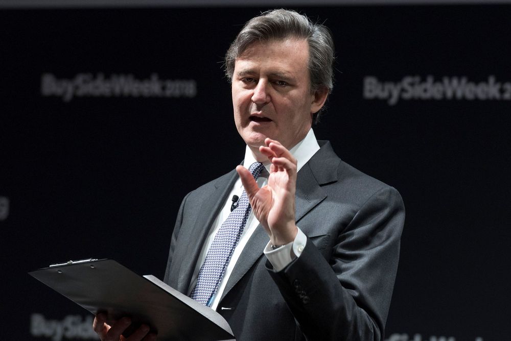 A man in a dark suit speaks at a BuySideWeek conference podium, gesturing with his hand.