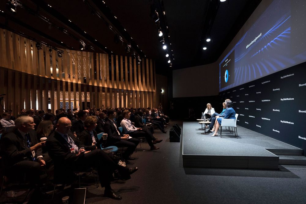Panel discussion on stage in modern auditorium with audience seated in rows under dramatic lighting.