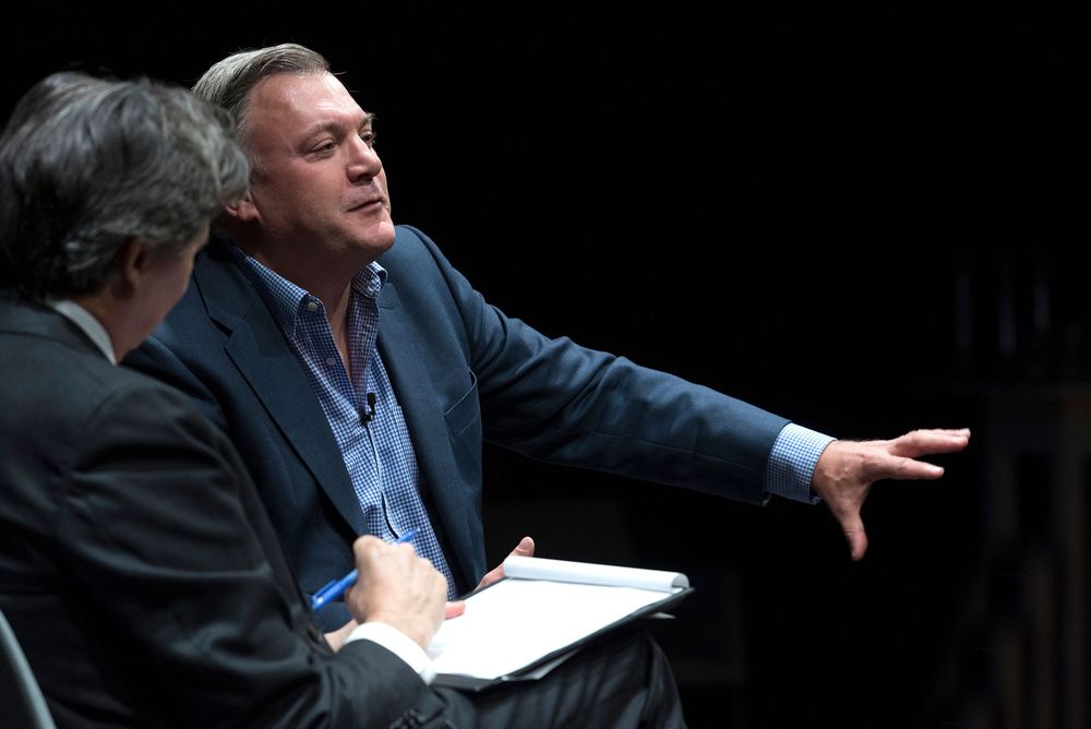 Two men in suits engaged in discussion on stage, one gesturing expressively while the other takes notes.