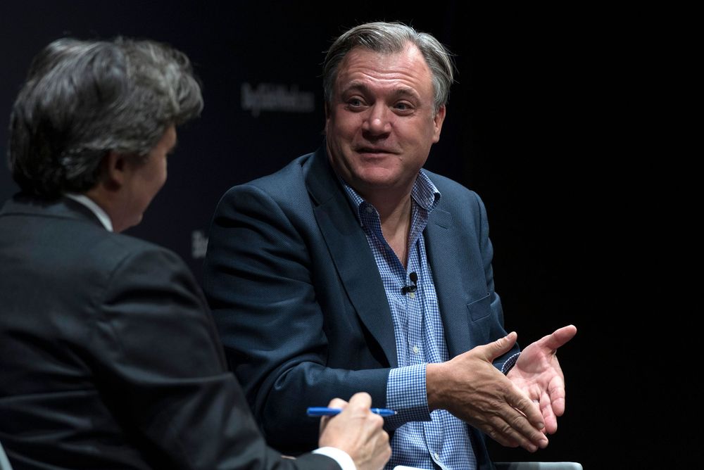 Two businessmen in suits having a conversation on stage, one smiling while the other listens attentively.
