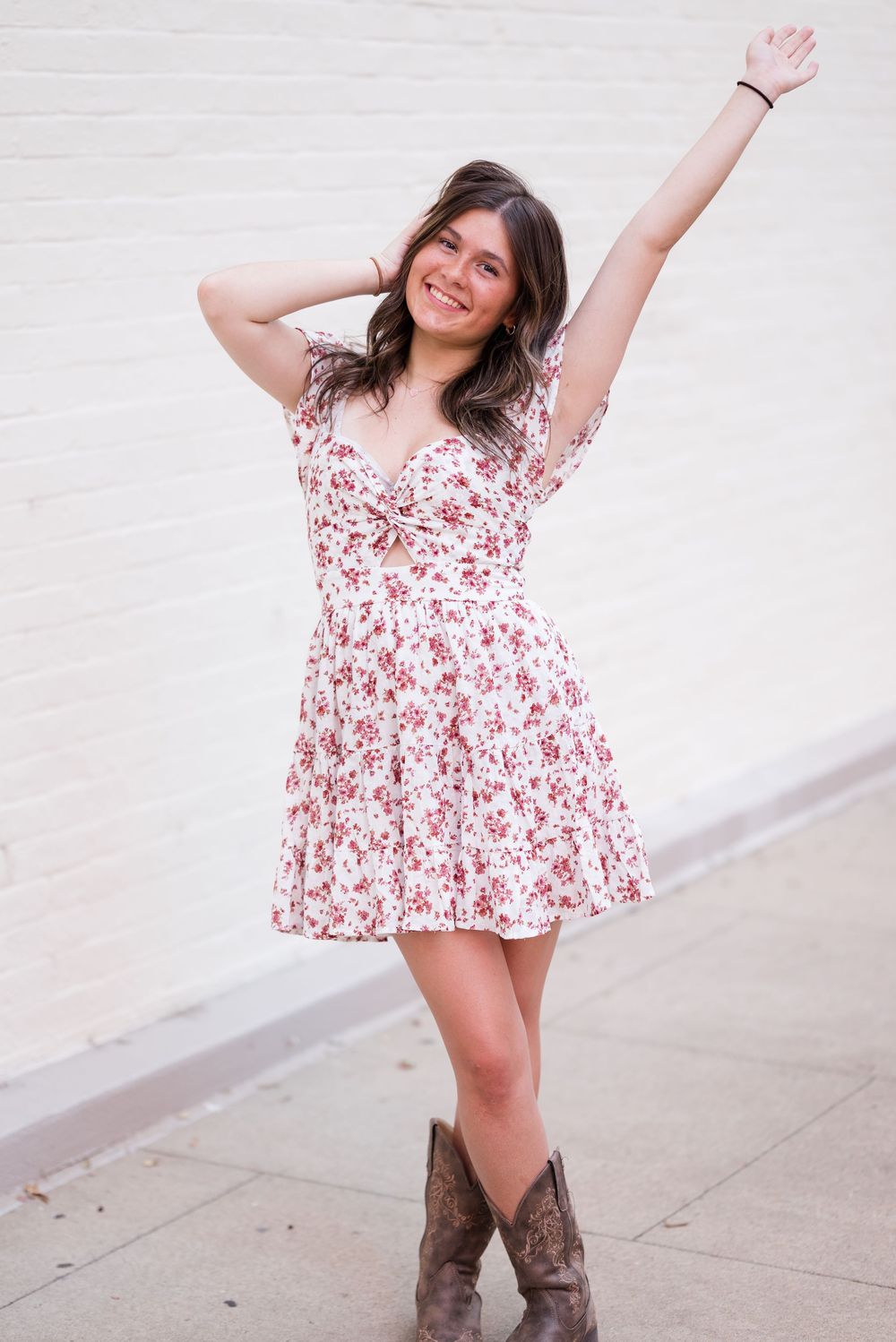 Playful poses in a white and red floral dress paired with brown cowboy boots against a white wall.