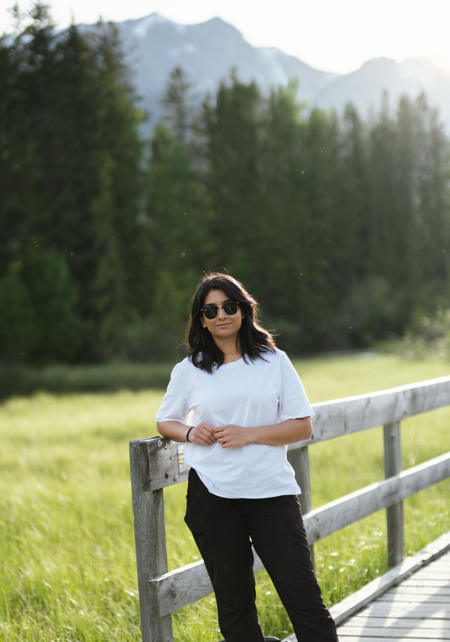 Person in white top and black pants poses along wooden fence with mountain backdrop and sunlit meadow.