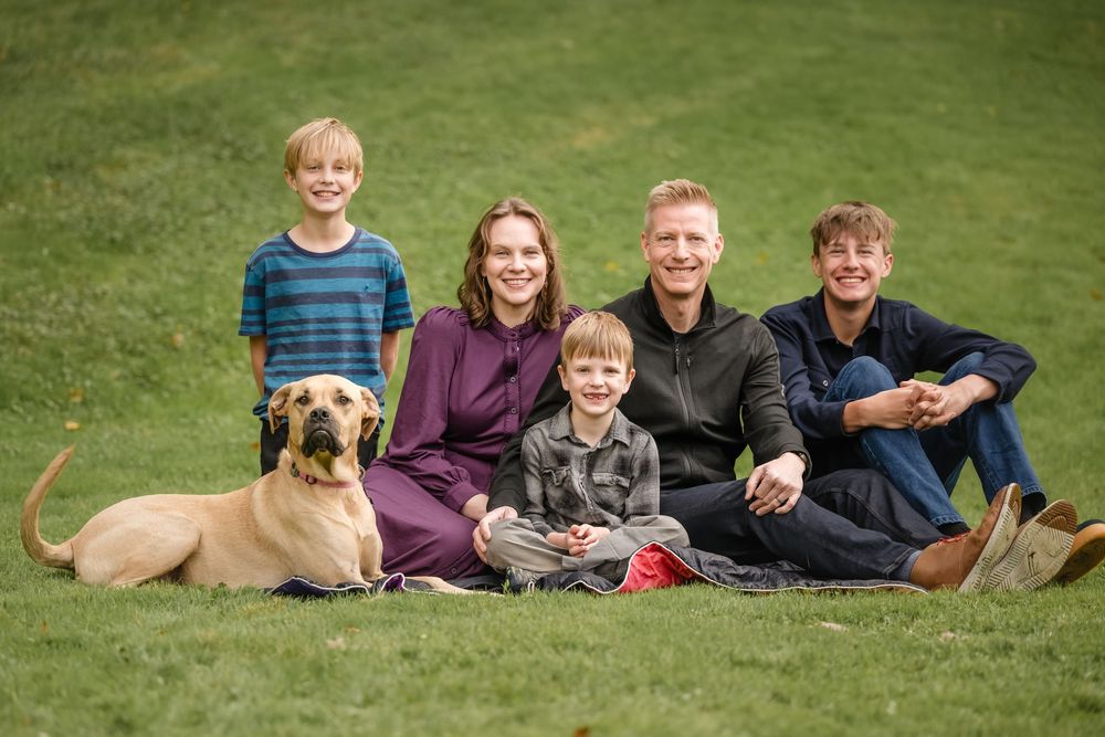 Happy family with three boys and a large tan dog sitting together on green grass outdoors.