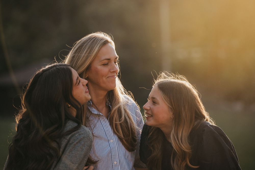 Three women smiling and laughing together outdoors in warm golden sunlight, sharing a joyful moment.