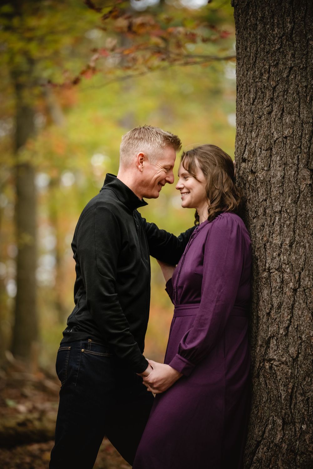 Couple sharing a romantic moment leaning against a tree in an autumn forest, woman in purple dress smiling.