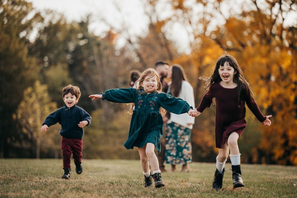 Three joyful children run through a grassy autumn park with colorful fall foliage trees in the background.
