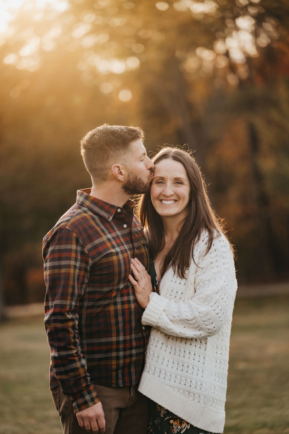 A couple shares a romantic moment outdoors in autumn, man kissing woman's cheek as she smiles in golden sunset light.