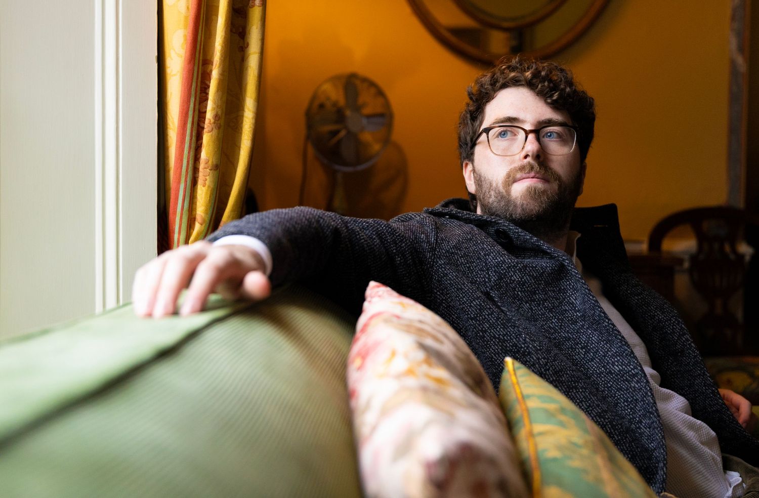 A person wearing glasses and a dark sweater reclines on a green couch in a warmly lit room with a fan visible in background.
