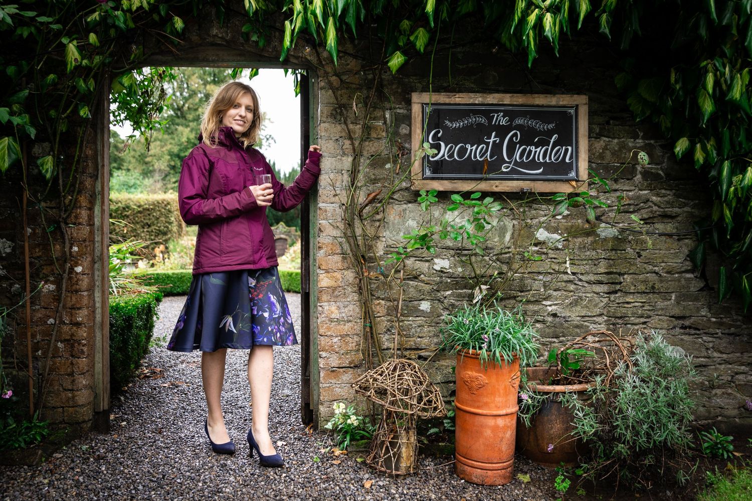 Standing by a rustic stone wall with a Secret Garden sign, holding an umbrella in a purple coat and skirt.
