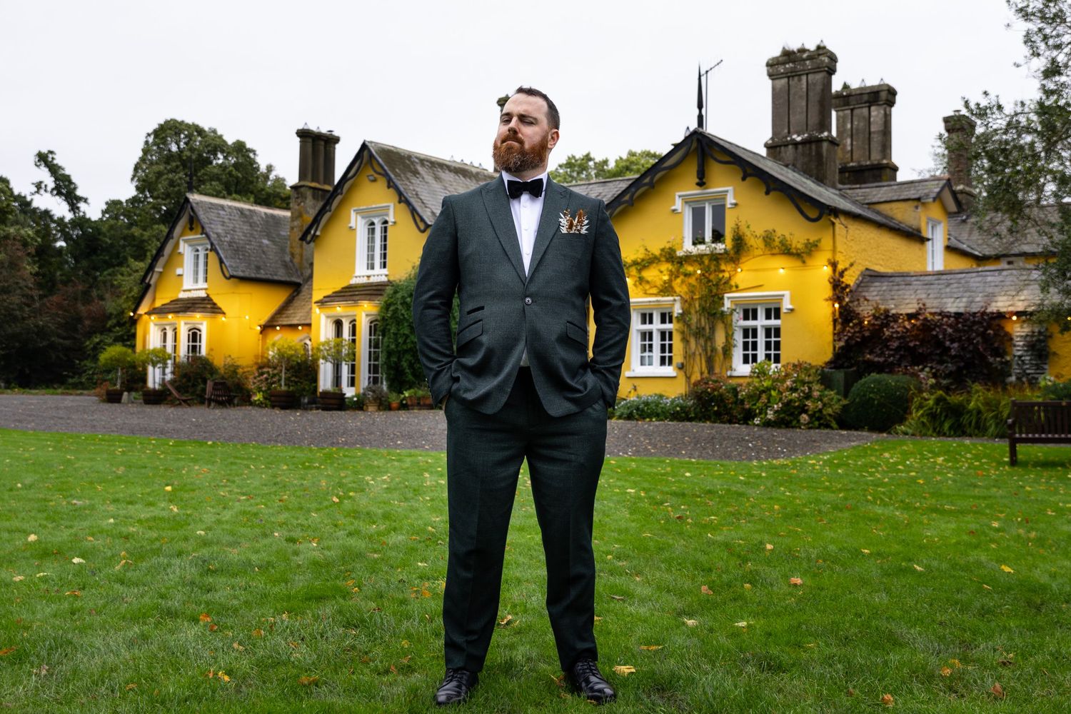 Figure in black tuxedo standing on lawn in front of yellow manor house with castle turret in Scotland.