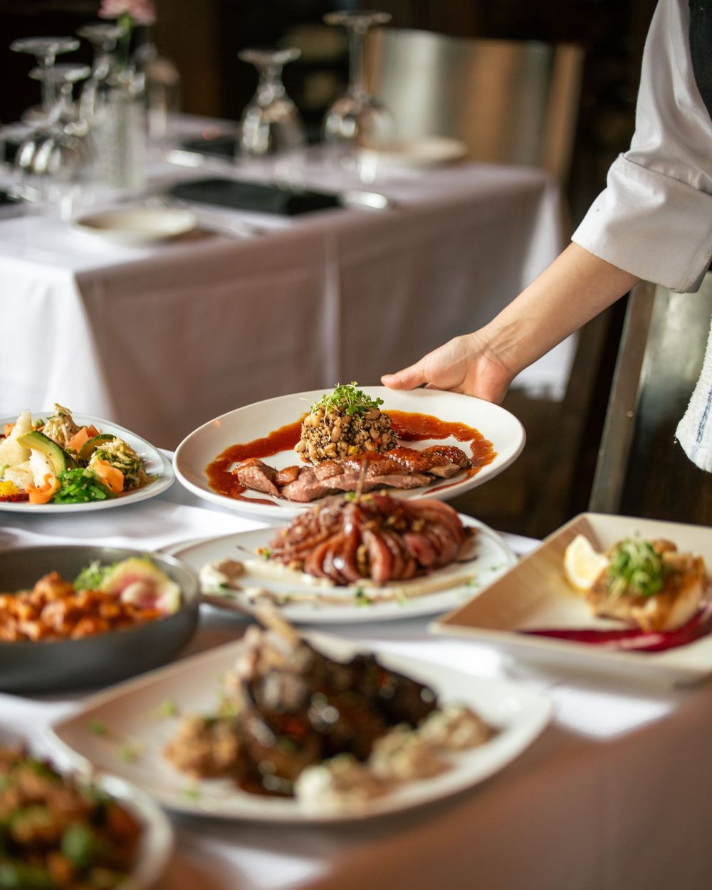 A server presenting a meat dish over a table filled with multiple gourmet plates at a restaurant.