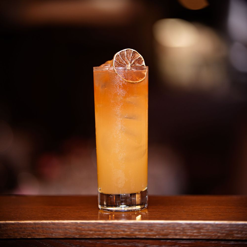 A tall amber cocktail with a dehydrated citrus wheel garnish in a highball glass on a wooden bar.