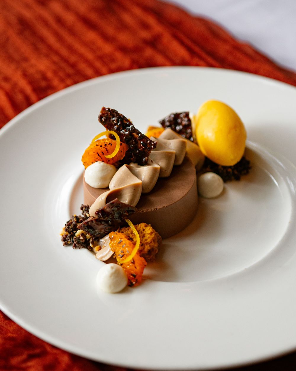 An artistic plated chocolate dessert with yellow sorbet, candied orange, and white meringue dots.