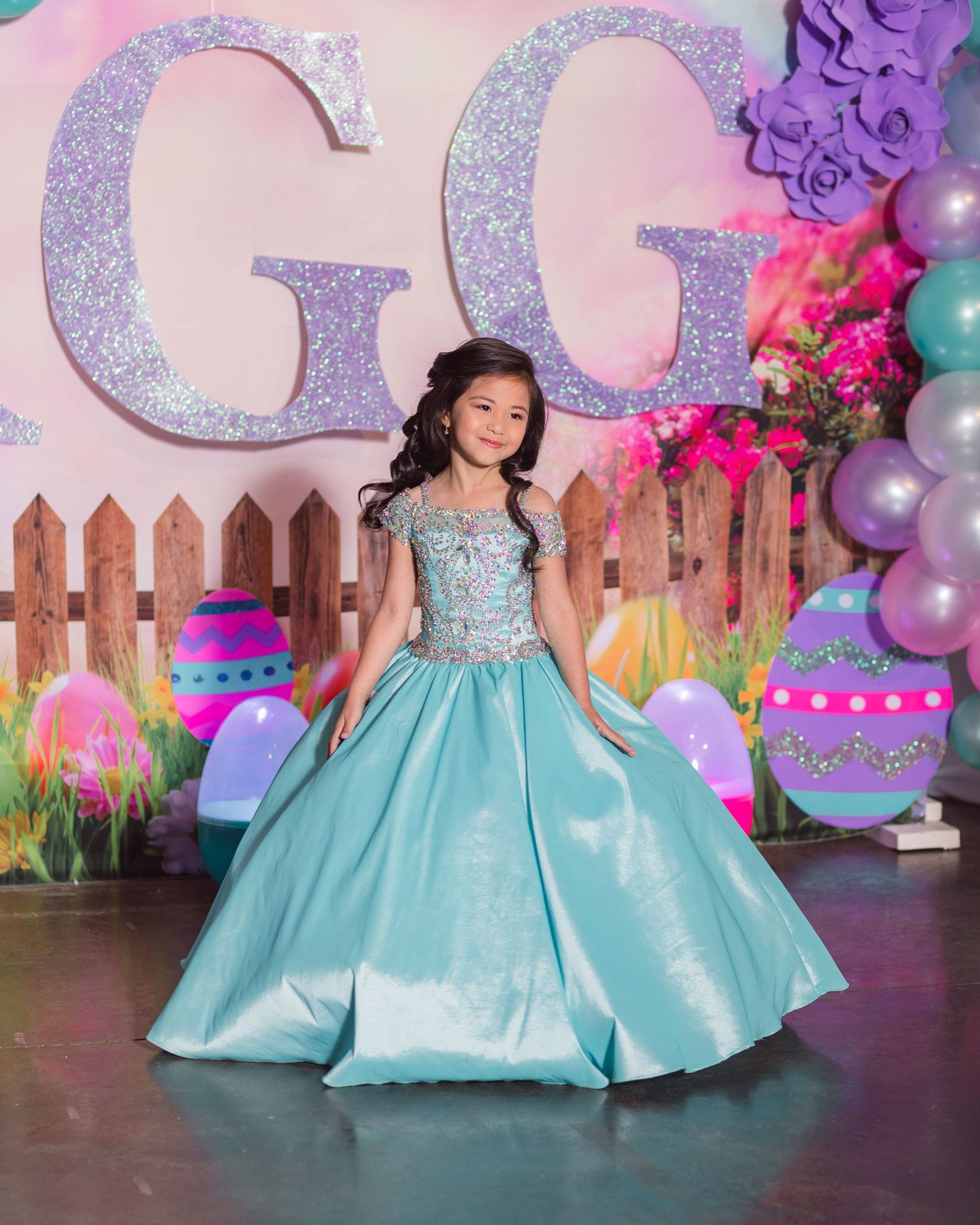 Young girl in turquoise ball gown poses at birthday party with glitter decorations and balloons.