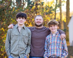 Three people standing together outdoors wearing casual clothing with fall foliage in the background.