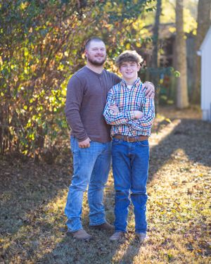 Two people standing together and smiling in front of autumn trees and a white building.