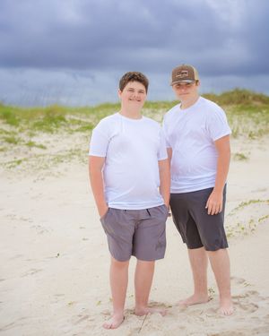 Two people in white shirts stand together on sandy beach with dunes behind them.