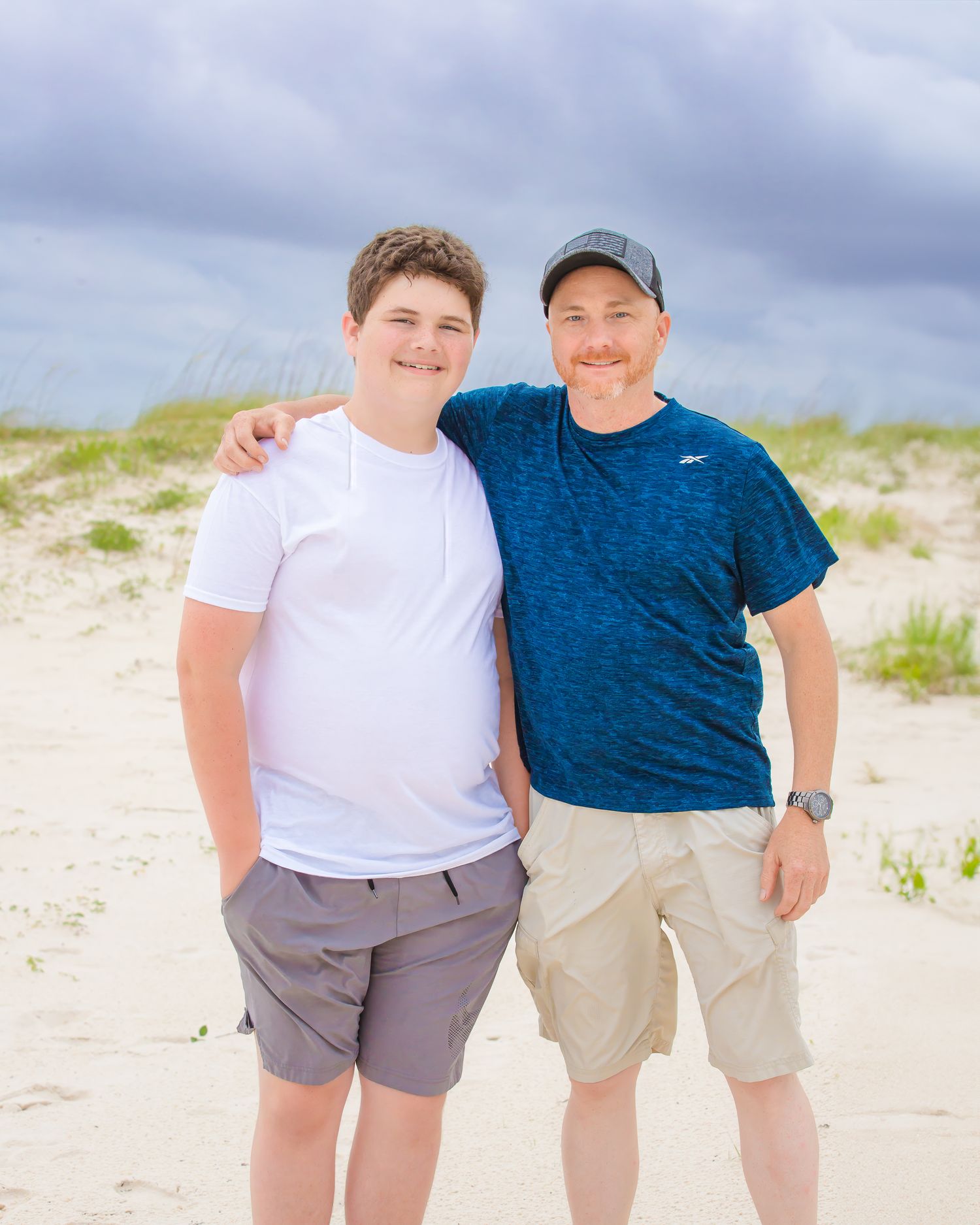 Two people pose together on beach, one in white shirt and one in blue shirt.