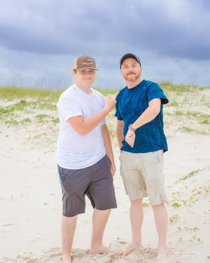Two people in casual attire pose on sandy beach with stormy sky background.