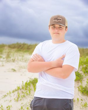Person in a white t-shirt and tan cap poses confidently on sandy dunes with cloudy sky in background.