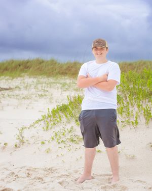Person in white t-shirt and cap stands barefoot on sandy beach dunes against stormy sky.