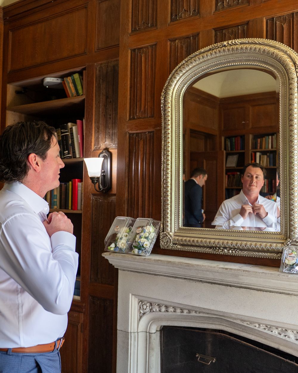 A man adjusts his collar while looking in an ornate gold mirror above a fireplace in a wood-paneled library room.