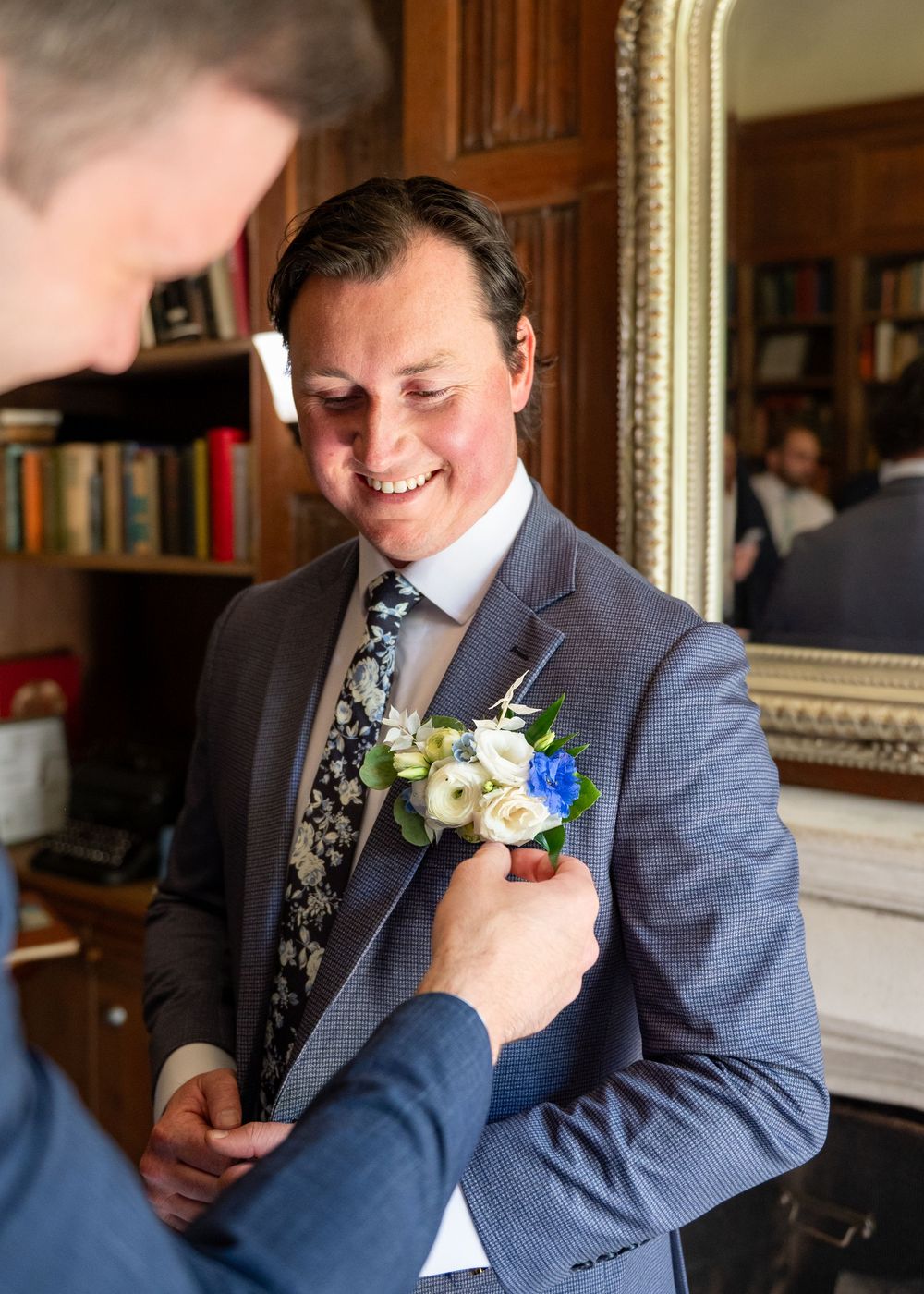 Smiling groom having a floral boutonniere pinned to his gray suit jacket in an elegant wood-paneled room.