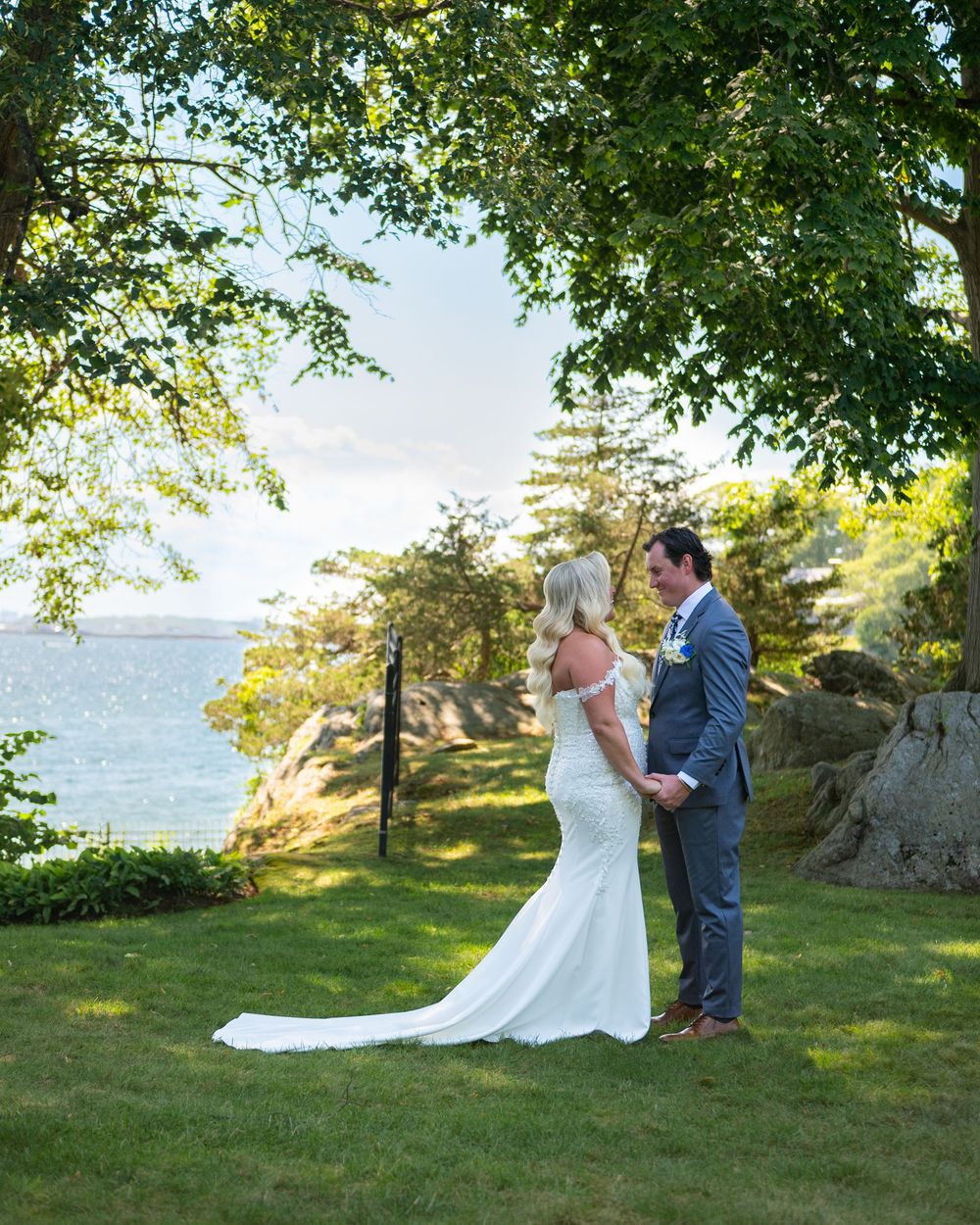 Bride and groom hold hands facing a scenic waterfront view, framed by lush green trees on a sunny wedding day.