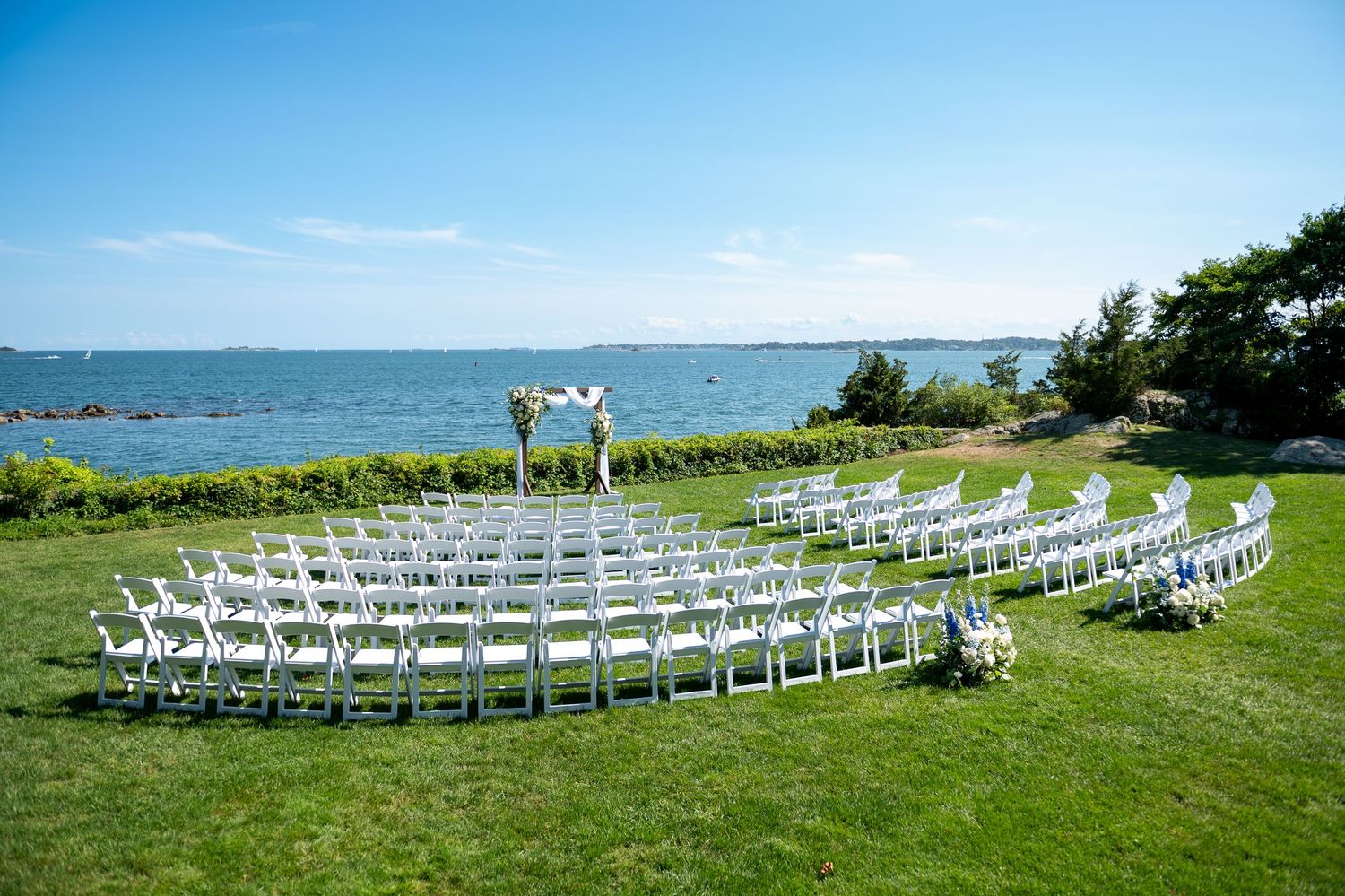 Outdoor waterfront wedding ceremony setup with white chairs arranged in semicircle on green lawn overlooking blue ocean.