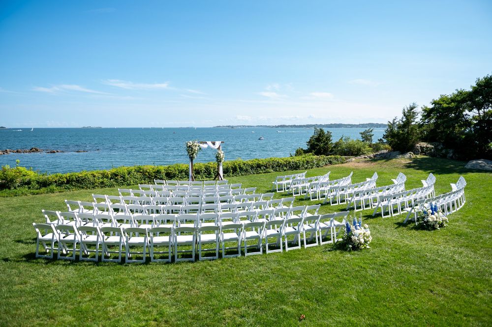 Outdoor waterfront wedding ceremony setup with white chairs arranged in semicircle on green lawn overlooking blue ocean.