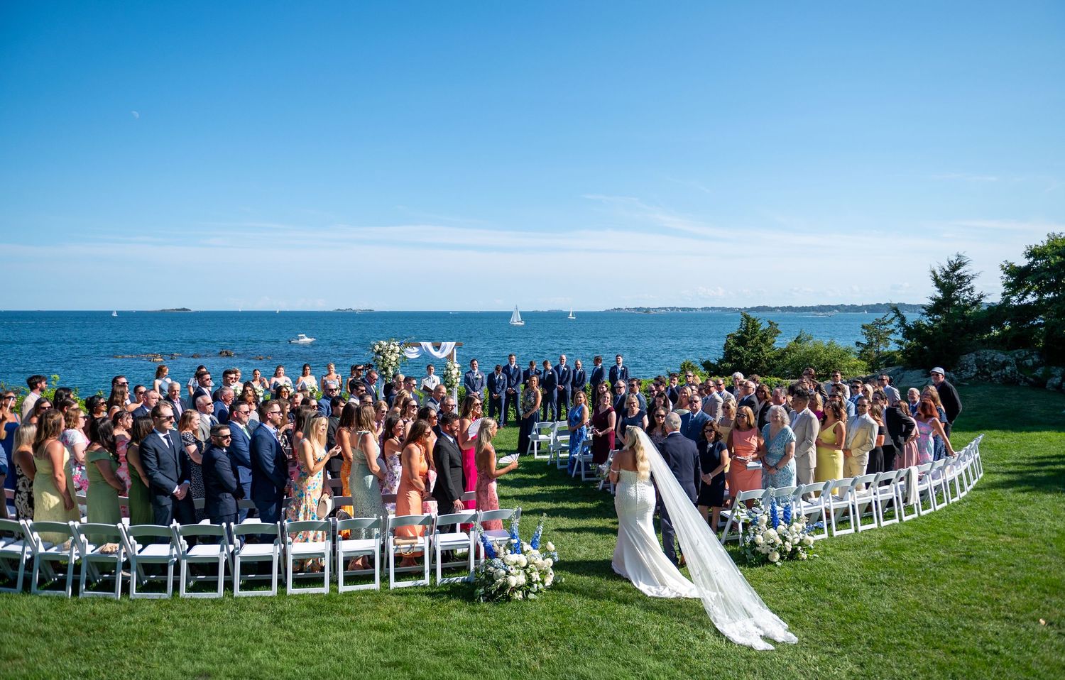 Outdoor wedding ceremony on a green lawn overlooking the ocean, with guests seated in a semicircle around the bride and groom.