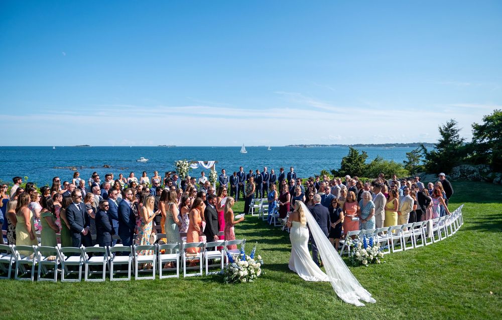 Outdoor wedding ceremony on a green lawn overlooking the ocean, with guests seated in a semicircle around the bride and groom.