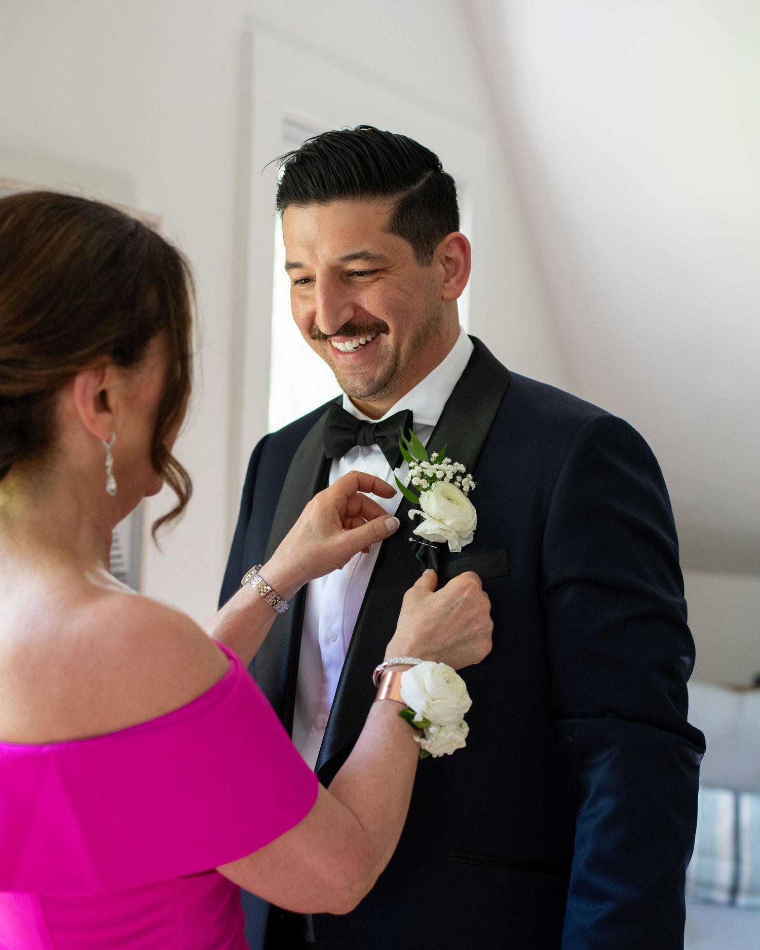 A smiling groom in a navy tuxedo gets his white boutonniere pinned by a woman in a pink off-shoulder dress.