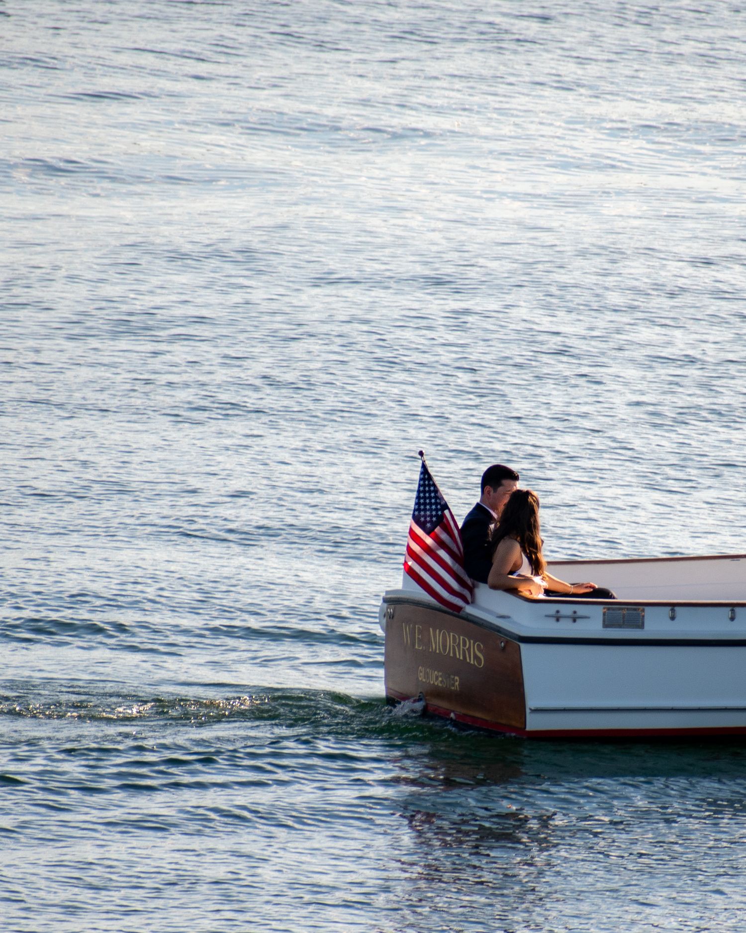Couple sharing a romantic kiss on a classic wooden boat with an American flag on calm blue water.