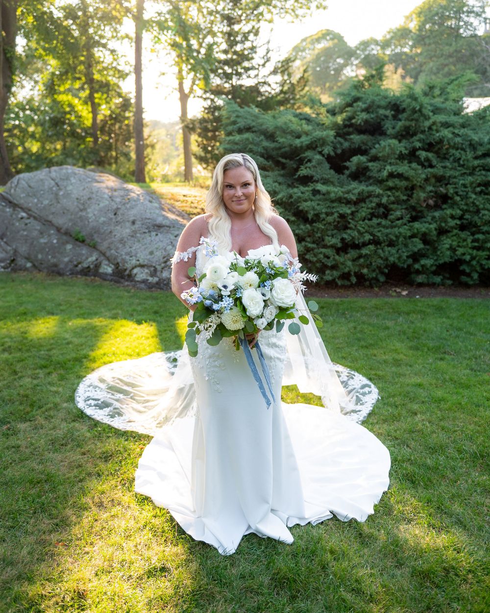 Bride in white off-shoulder gown holding white floral bouquet stands on green lawn with trees and rocks backdrop.