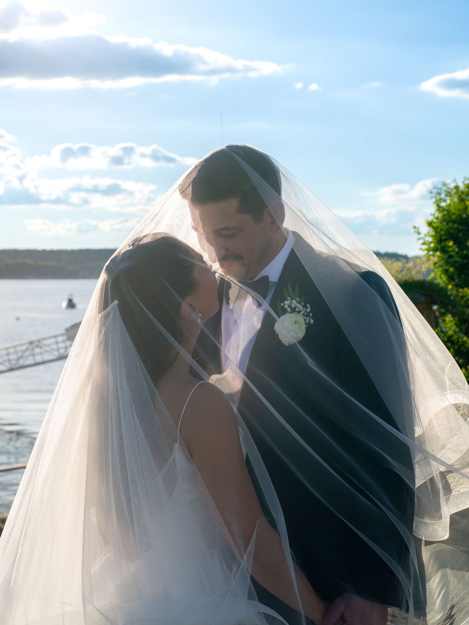 Bride and groom share an intimate moment under a flowing wedding veil with a scenic waterfront backdrop on their wedding day.