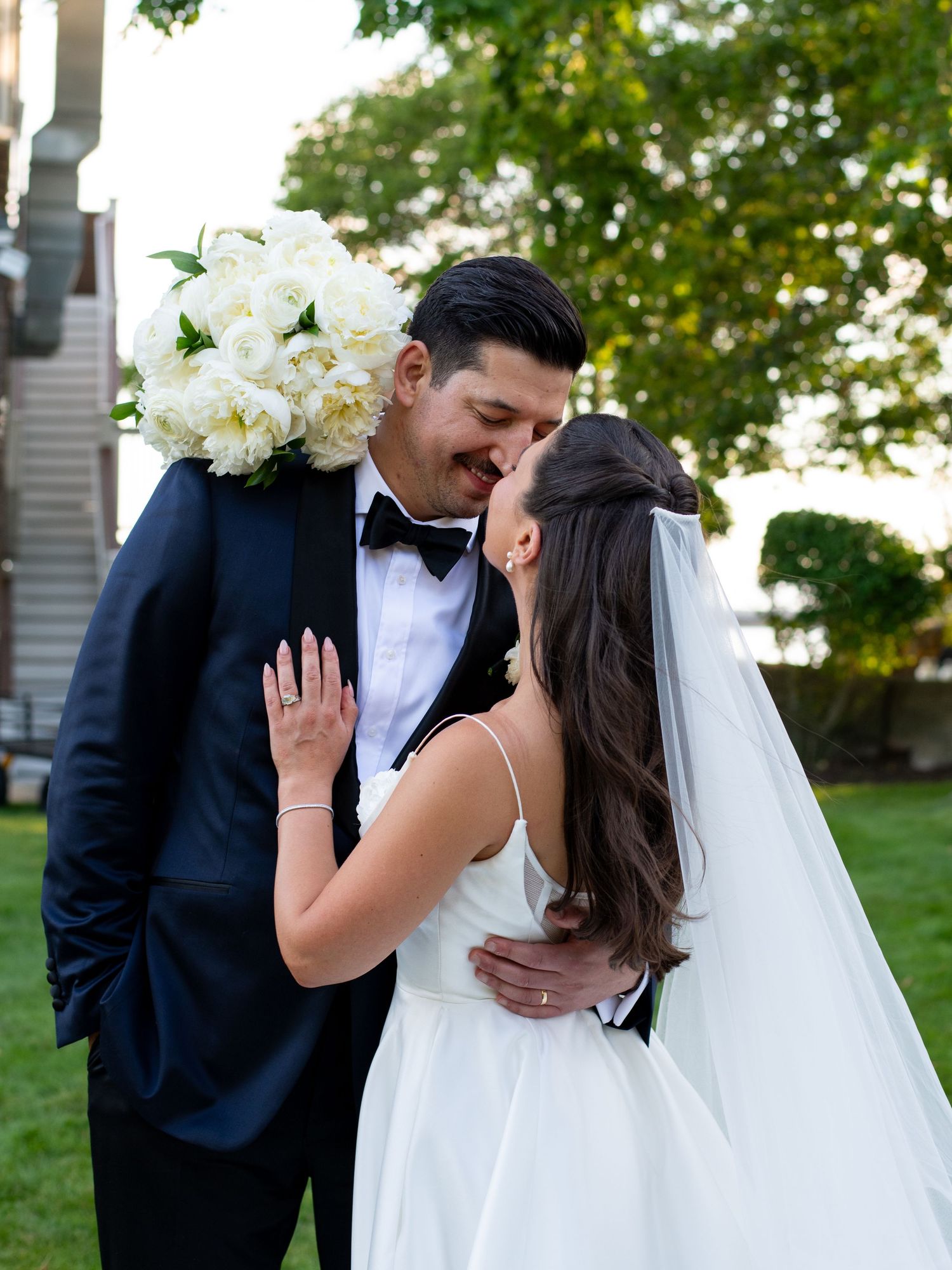 Bride and groom sharing a romantic kiss outdoors, bride holding white peony bouquet, groom in navy tuxedo.