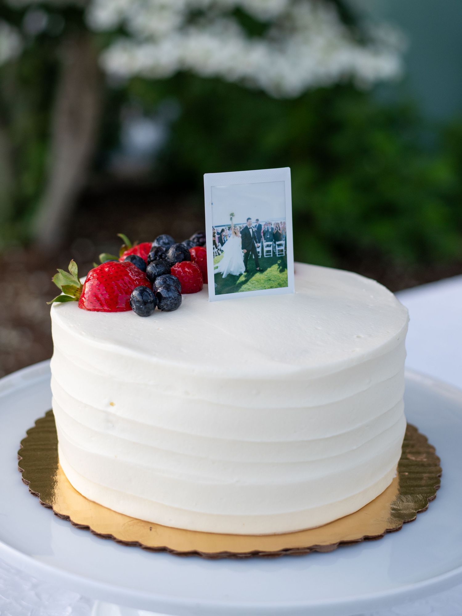 Elegant white wedding cake with fresh berries and a photo topper on a gold cake board.