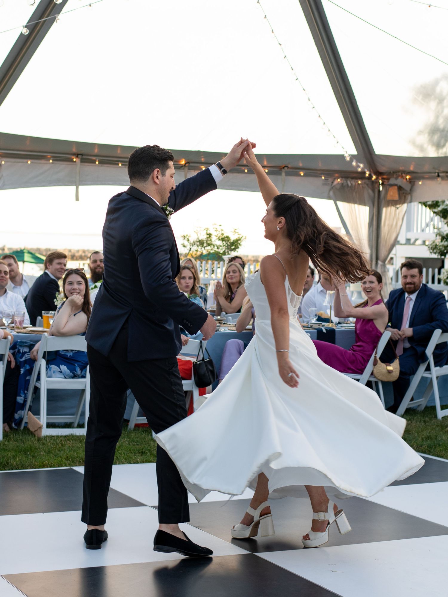 Bride and groom share first dance under outdoor tent at wedding reception, guests watching as bride twirls in white gown.