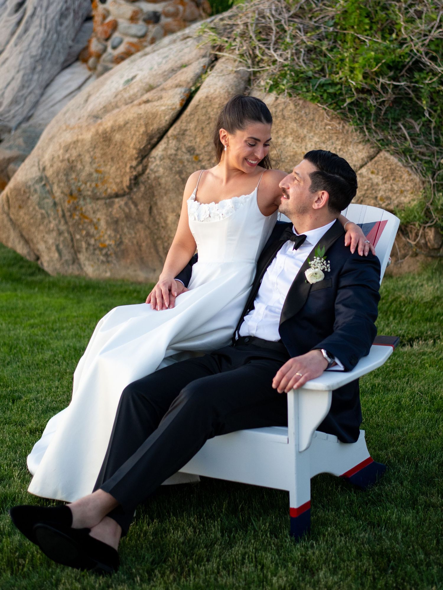 Bride in white gown sits on groom's lap in white Adirondack chair, sharing a tender moment outdoors.