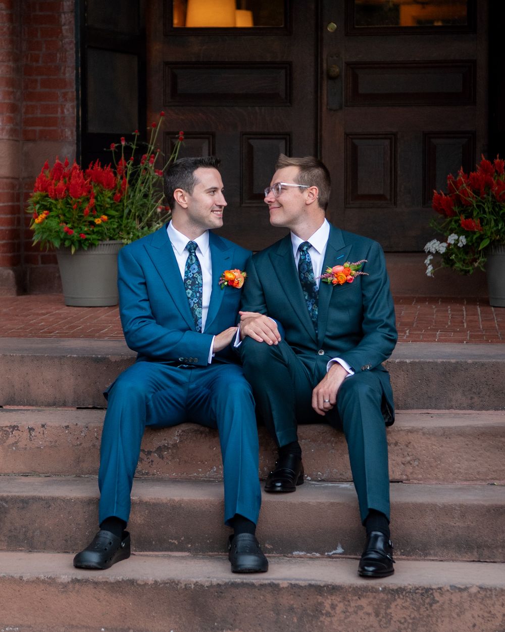Two grooms in teal suits with floral boutonnieres sit on brick steps, smiling at each other on their wedding day.