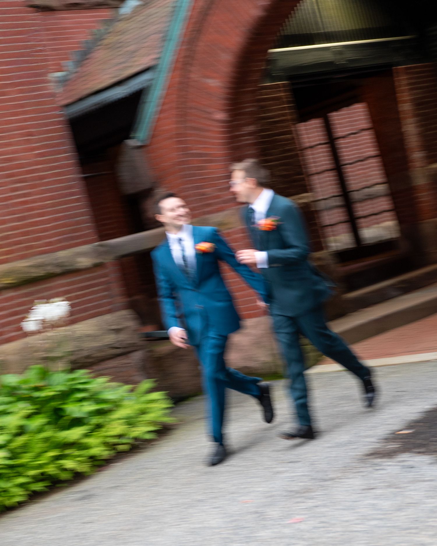 Two grooms in blue suits laugh and walk together outside a brick building on their wedding day.