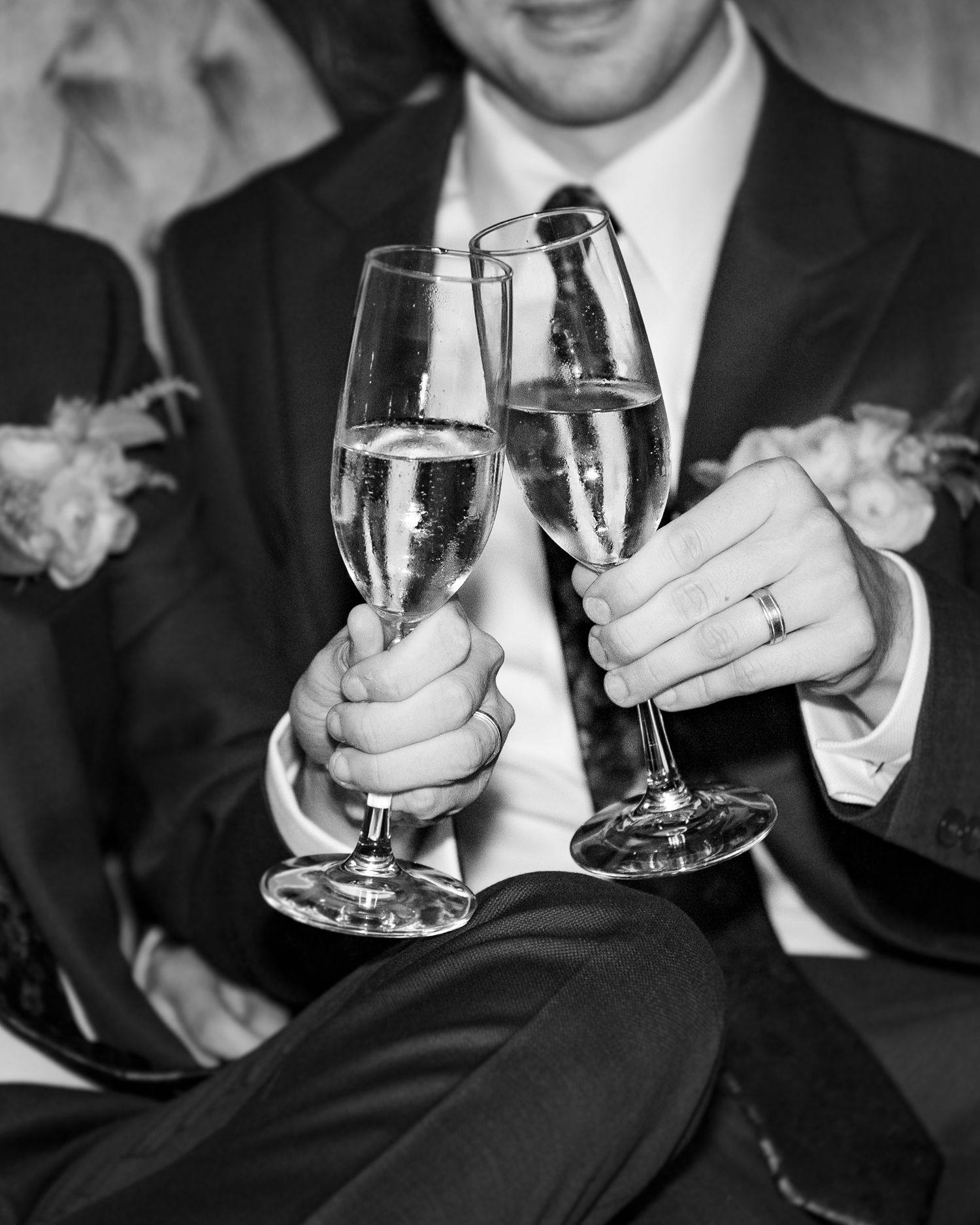 Black and white photo of newlyweds toasting with champagne flutes, wedding rings visible, elegant formal attire.